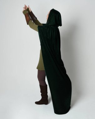 Kızıl saçlı kadın modelin tam boy portresi, yeşil ortaçağ fantezi kostümü, uzun kadife kapüşonlu pelerin. Duruş, geri geri yürüme, izole beyaz stüdyo arka planı.