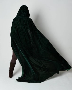 Kızıl saçlı kadın modelin tam boy portresi, yeşil ortaçağ fantezi kostümü, uzun kadife kapüşonlu pelerin. Duruş, geri geri yürüme, izole beyaz stüdyo arka planı.