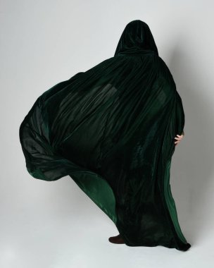 Kızıl saçlı kadın modelin tam boy portresi, yeşil ortaçağ fantezi kostümü, uzun kadife kapüşonlu pelerin. Duruş, geri geri yürüme, izole beyaz stüdyo arka planı.