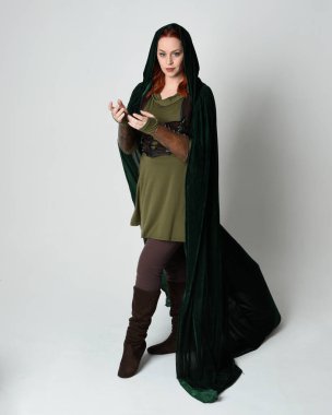 Yeşil ortaçağ fantezi deri zırh giyen kızıl saçlı kadın modelin tam boy portresi, dramatik uzun kadife kapüşonlu pelerin. Yürüyen poz, izole edilmiş beyaz stüdyo arka planı..