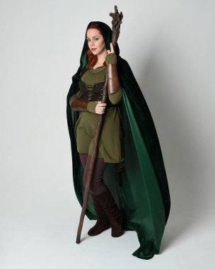 Yeşil ortaçağ fantazisi deri zırh giyen kızıl saçlı kadın modelin tam boy portresi, çarpıcı uzun, kadife kapüşonlu pelerin, ahşap büyücü değneği tutan. Yürüyen poz, izole edilmiş beyaz stüdyo arka planı..