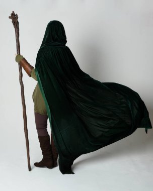 Yeşil ortaçağ fantazisi deri zırh giyen kızıl saçlı kadın modelin tam boy portresi, çarpıcı uzun, kadife kapüşonlu pelerin, ahşap büyücü değneği tutan. Yürüyen poz, izole edilmiş beyaz stüdyo arka planı..