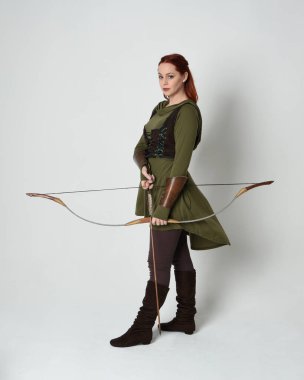 Yeşil ortaçağ fantezi kostümü giyen kızıl saçlı kadın modelin tam boy portresi. Okçuluk yayı ve ok tabancası tutan, beyaz stüdyo arka planında izole edilmiş bir şekilde duran..
