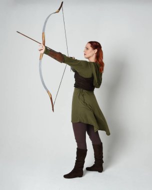 Yeşil ortaçağ fantezi kostümü giyen kızıl saçlı kadın modelin tam boy portresi. Okçuluk yayı ve ok tabancası tutan, beyaz stüdyo arka planında izole edilmiş bir şekilde duran..