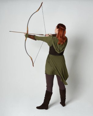 Yeşil ortaçağ fantezi kostümü giyen kızıl saçlı kadın modelin tam boy portresi. Okçuluk yayı ve ok tabancası tutan, beyaz stüdyo arka planında izole edilmiş bir şekilde duran..