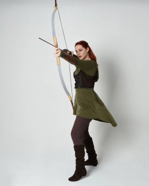 Yeşil ortaçağ fantezi kostümü giyen kızıl saçlı kadın modelin tam boy portresi. Okçuluk yayı ve ok tabancası tutan, beyaz stüdyo arka planında izole edilmiş bir şekilde duran..