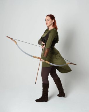 Yeşil ortaçağ fantezi kostümü giyen kızıl saçlı kadın modelin tam boy portresi. Okçuluk yayı ve ok tabancası tutan, beyaz stüdyo arka planında izole edilmiş bir şekilde duran..