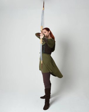 Yeşil ortaçağ fantezi kostümü giyen kızıl saçlı kadın modelin tam boy portresi. Okçuluk yayı ve ok tabancası tutan, beyaz stüdyo arka planında izole edilmiş bir şekilde duran..