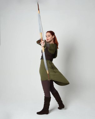 Yeşil ortaçağ fantezi kostümü giyen kızıl saçlı kadın modelin tam boy portresi. Okçuluk yayı ve ok tabancası tutan, beyaz stüdyo arka planında izole edilmiş bir şekilde duran..