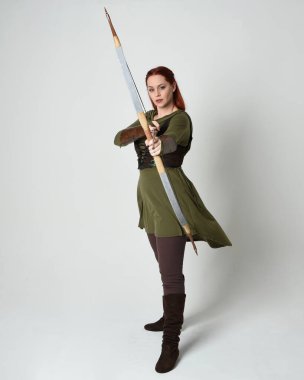 Yeşil ortaçağ fantezi kostümü giyen kızıl saçlı kadın modelin tam boy portresi. Okçuluk yayı ve ok tabancası tutan, beyaz stüdyo arka planında izole edilmiş bir şekilde duran..