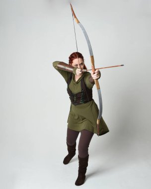 Yeşil ortaçağ fantezi kostümü giyen kızıl saçlı kadın modelin tam boy portresi. Okçuluk yayı ve ok tabancası tutan, beyaz stüdyo arka planında izole edilmiş bir şekilde duran..