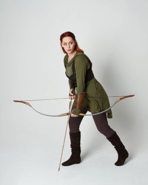 Yeşil ortaçağ fantezi kostümü giyen kızıl saçlı kadın modelin tam boy portresi. Okçuluk yayı ve ok tabancası tutan, beyaz stüdyo arka planında izole edilmiş bir şekilde duran..