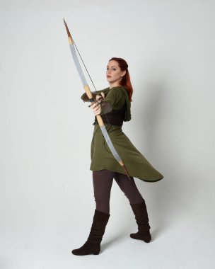 Yeşil ortaçağ fantezi kostümü giyen kızıl saçlı kadın modelin tam boy portresi. Okçuluk yayı ve ok tabancası tutan, beyaz stüdyo arka planında izole edilmiş bir şekilde duran..