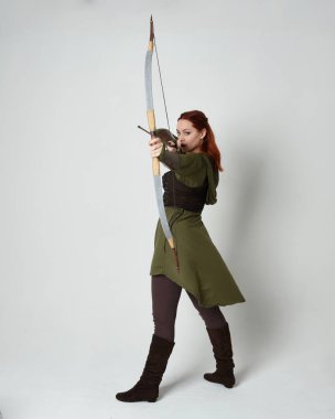 Yeşil ortaçağ fantezi kostümü giyen kızıl saçlı kadın modelin tam boy portresi. Okçuluk yayı ve ok tabancası tutan, beyaz stüdyo arka planında izole edilmiş bir şekilde duran..