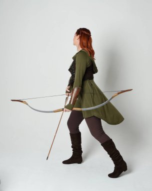 Yeşil ortaçağ fantezi kostümü giyen kızıl saçlı kadın modelin tam boy portresi. Okçuluk yayı ve ok tabancası tutan, beyaz stüdyo arka planında izole edilmiş bir şekilde duran..