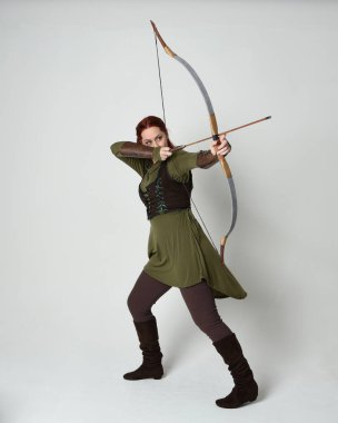 Yeşil ortaçağ fantezi kostümü giyen kızıl saçlı kadın modelin tam boy portresi. Okçuluk yayı ve ok tabancası tutan, beyaz stüdyo arka planında izole edilmiş bir şekilde duran..