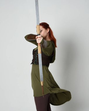 Yeşil ortaçağ fantezi kostümü giyen kızıl saçlı kadın modelin yakın plan portresi. Okçuluk yayı ve ok tabancası tutan, beyaz stüdyo arka planında izole edilmiş bir şekilde duran.