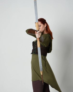 Yeşil ortaçağ fantezi kostümü giyen kızıl saçlı kadın modelin yakın plan portresi. Okçuluk yayı ve ok tabancası tutan, beyaz stüdyo arka planında izole edilmiş bir şekilde duran.