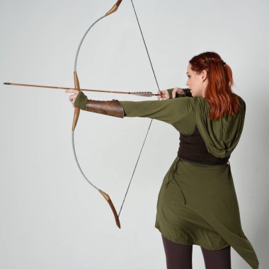 Yeşil ortaçağ fantezi kostümü giyen kızıl saçlı kadın modelin yakın plan portresi. Okçuluk yayı ve ok tabancası tutan, beyaz stüdyo arka planında izole edilmiş bir şekilde duran.