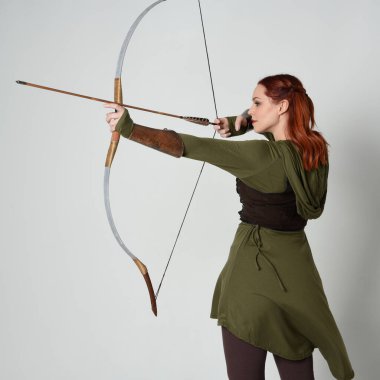 Yeşil ortaçağ fantezi kostümü giyen kızıl saçlı kadın modelin yakın plan portresi. Okçuluk yayı ve ok tabancası tutan, beyaz stüdyo arka planında izole edilmiş bir şekilde duran.