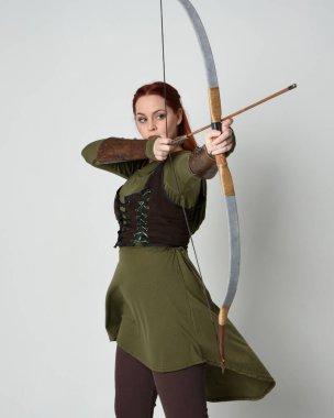 Yeşil ortaçağ fantezi kostümü giyen kızıl saçlı kadın modelin yakın plan portresi. Okçuluk yayı ve ok tabancası tutan, beyaz stüdyo arka planında izole edilmiş bir şekilde duran.