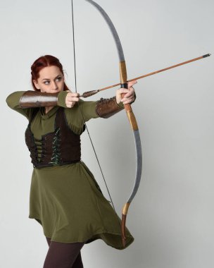 Yeşil ortaçağ fantezi kostümü giyen kızıl saçlı kadın modelin yakın plan portresi. Okçuluk yayı ve ok tabancası tutan, beyaz stüdyo arka planında izole edilmiş bir şekilde duran.