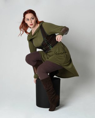 Yeşil ortaçağ fantezi kostümü giyen kızıl saçlı kadın modelinin tam boy portresi. Deri cephanelik. Beyaz stüdyo arka planında izole edilmiş, kambur bir poz..