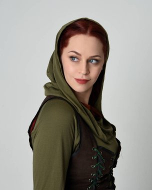 Yeşil ve kahverengi ortaçağ fantezi kostümü, kapüşonlu gömlek ve kahverengi tunik giyen güzel kızıl saçlı kadın modelin yakın portresi. Beyaz stüdyoda izole 