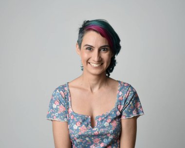 Alternatif kadın modelin yakın plan portresi, pembe ve gri saç kesimi tarzıyla, çiçekli mavi bir bluz giyerek. Etkileyici bir yüz ifadesi. Stüdyo arka planında izole.