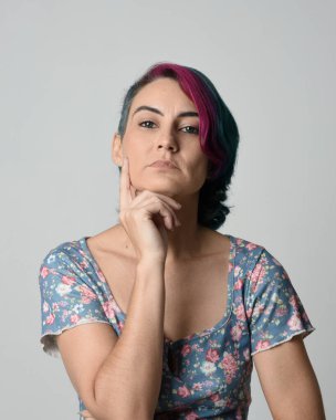 Alternatif kadın modelin yakın plan portresi, pembe ve gri saç kesimi tarzıyla, çiçekli mavi bir bluz giyerek. Etkileyici bir yüz ifadesi. Stüdyo arka planında izole.