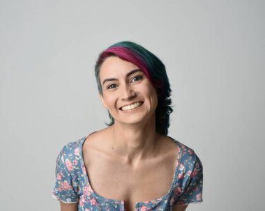 Alternatif kadın modelin yakın plan portresi, pembe ve gri saç kesimi tarzıyla, çiçekli mavi bir bluz giyerek. Etkileyici bir yüz ifadesi. Stüdyo arka planında izole.