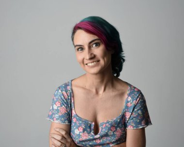 Alternatif kadın modelin yakın plan portresi, pembe ve gri saç kesimi tarzıyla, çiçekli mavi bir bluz giyerek. Etkileyici bir yüz ifadesi. Stüdyo arka planında izole.