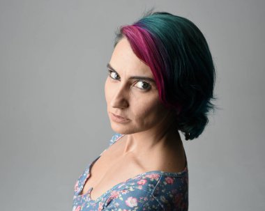 Alternatif kadın modelin yakın plan portresi, pembe ve gri saç kesimi tarzıyla, çiçekli mavi bir bluz giyerek. Etkileyici bir yüz ifadesi. Stüdyo arka planında izole.