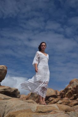 Beyaz tanrıça elbisesi giymiş kadın modelin portresi kayalık okyanus kıyısının kayalık uçurumlarla çevrili dramatik doğal manzarası. Castle Rock, Busselton, Batı Avustralya