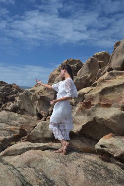 Beyaz tanrıça elbisesi giymiş kadın modelin portresi kayalık okyanus kıyısının kayalık uçurumlarla çevrili dramatik doğal manzarası. Castle Rock, Busselton, Batı Avustralya