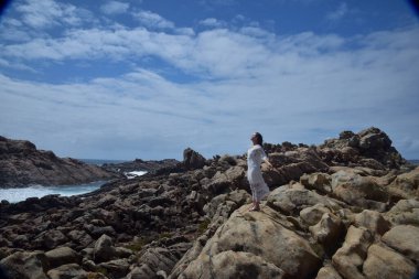 Beyaz tanrıça elbisesi giymiş kadın modelin portresi kayalık okyanus kıyısının kayalık uçurumlarla çevrili dramatik doğal manzarası. Castle Rock, Busselton, Batı Avustralya