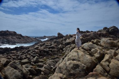 Beyaz tanrıça elbisesi giymiş kadın modelin portresi kayalık okyanus kıyısının kayalık uçurumlarla çevrili dramatik doğal manzarası. Castle Rock, Busselton, Batı Avustralya