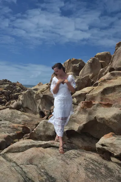 Beyaz tanrıça elbisesi giymiş kadın modelin portresi kayalık okyanus kıyısının kayalık uçurumlarla çevrili dramatik doğal manzarası. Castle Rock, Busselton, Batı Avustralya