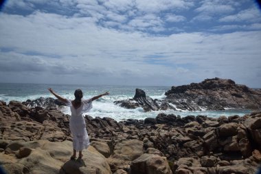 Beyaz tanrıça elbisesi giymiş kadın modelin portresi kayalık okyanus kıyısının kayalık uçurumlarla çevrili dramatik doğal manzarası. Castle Rock, Busselton, Batı Avustralya