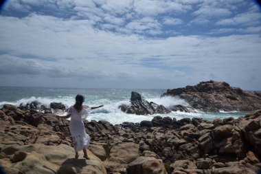 Beyaz tanrıça elbisesi giymiş kadın modelin portresi kayalık okyanus kıyısının kayalık uçurumlarla çevrili dramatik doğal manzarası. Castle Rock, Busselton, Batı Avustralya