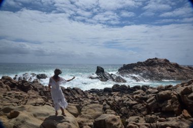 Beyaz tanrıça elbisesi giymiş kadın modelin portresi kayalık okyanus kıyısının kayalık uçurumlarla çevrili dramatik doğal manzarası. Castle Rock, Busselton, Batı Avustralya