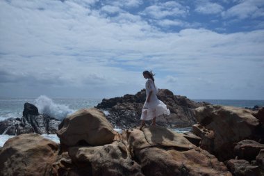 Beyaz tanrıça elbisesi giymiş kadın modelin portresi kayalık okyanus kıyısının kayalık uçurumlarla çevrili dramatik doğal manzarası. Castle Rock, Busselton, Batı Avustralya