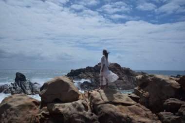 Beyaz tanrıça elbisesi giymiş kadın modelin portresi kayalık okyanus kıyısının kayalık uçurumlarla çevrili dramatik doğal manzarası. Castle Rock, Busselton, Batı Avustralya