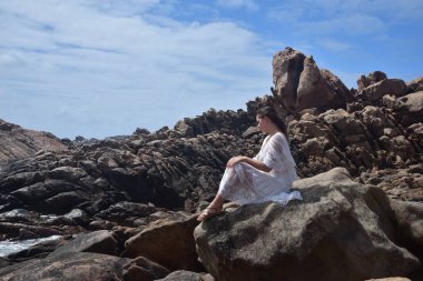 Beyaz tanrıça elbisesi giymiş kadın modelin portresi kayalık okyanus kıyısının kayalık uçurumlarla çevrili dramatik doğal manzarası. Castle Rock, Busselton, Batı Avustralya