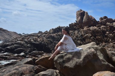 Beyaz tanrıça elbisesi giymiş kadın modelin portresi kayalık okyanus kıyısının kayalık uçurumlarla çevrili dramatik doğal manzarası. Castle Rock, Busselton, Batı Avustralya