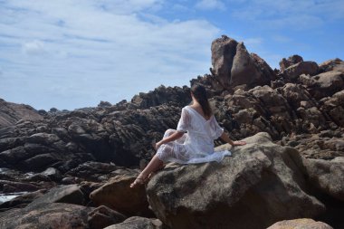 Beyaz tanrıça elbisesi giymiş kadın modelin portresi kayalık okyanus kıyısının kayalık uçurumlarla çevrili dramatik doğal manzarası. Castle Rock, Busselton, Batı Avustralya