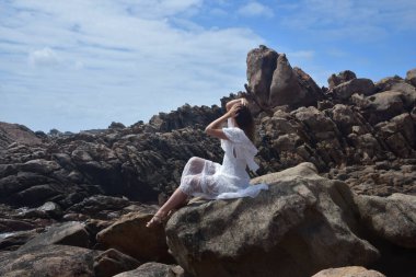 Beyaz tanrıça elbisesi giymiş kadın modelin portresi kayalık okyanus kıyısının kayalık uçurumlarla çevrili dramatik doğal manzarası. Castle Rock, Busselton, Batı Avustralya