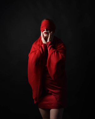 Kırmızı ipek elbise giyen ve gözlerini bağlayan kumaştan bir kadın manken portresi. Karanlık bir stüdyonun arka planında karamsar duygusal kol hareketleriyle izole edilmiş..