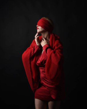 Kırmızı ipek elbise giyen ve gözlerini bağlayan kumaştan bir kadın manken portresi. Karanlık bir stüdyonun arka planında karamsar duygusal kol hareketleriyle izole edilmiş..