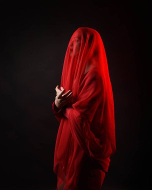 Dramatik bir portre. Kırmızı kumaş peçe takan, karamsar duygusal hareketleriyle karanlık bir stüdyo arka planında izole edilmiş hayalet bir kadın model..
